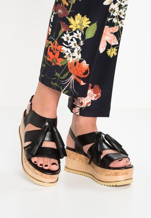 Fødder iført sorte læder sandaler med tofs og kork kilehæle, kombineret med sorte bukser med farverigt blomsterprint.