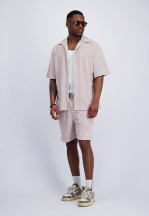 Mann trägt beige kurzärmliges Hemd mit Knöpfen, passende Shorts, weißes Tanktop, Sonnenbrille, hohe weiße Socken und Turnschuhe, steht vor einfachem Hintergrund.