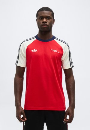 Mand iført rød og hvid kortærmet sportstrøje med marineblå krave og logoer på brystet samt stribede ærmer, stående mod en hvid baggrund.