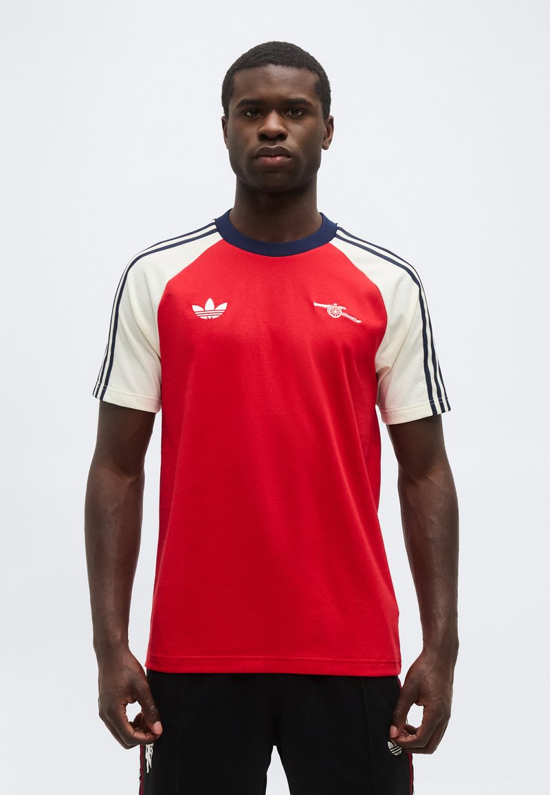 Άνδρας που φοράει κόκκινο και άσπρο αθλητικό κοντομάνικο πουκάμισο με σκούρο μπλε γιακά και λογότυπα στο στήθος και ριγέ μανίκια, στέκεται μπροστά σε λευκό φόντο.