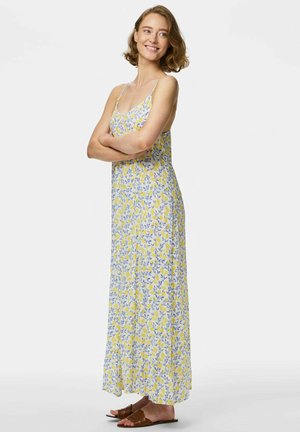 Giovane donna sorridente con le braccia incrociate, che indossa un vestito lungo senza maniche giallo e blu a fiori e sandali bassi marroni.