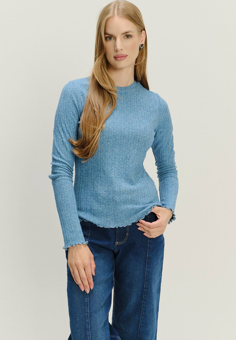 Maglione azzurro chiaro a coste con scollo rotondo e maniche lunghe, caratterizzato da un orlo ondulato e un motivo testurizzato. Indossato con pantaloni blu scuro.