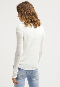 Pull en tricot blanc avec des manches longues, tissu texturé et coupe décontractée. Associé à un jean bleu clair ajusté. Vue de dos.