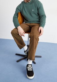 Sweat-shirt vert, pantalon marron avec coutures visibles, baskets noires avec lacets blancs, et chaussettes blanches, assis sur une chaise en bois.