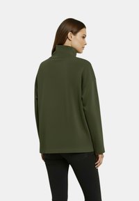 Maglione verde scuro con collo alto, maniche lunghe e vestibilità ampia. Tessuto liscio e design minimale. Indossato con jeans neri.