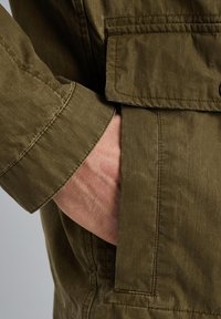 Veste olive en tissu texturé, avec des coutures visibles et une poche latérale où une main est placée à l'intérieur pour donner une idée d'échelle.
