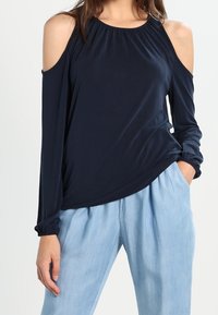 Tmavě modrý top s odhalenými rameny, dlouhými rukávy, nařaseným výstřihem a elastickými manžetami, doplněný světlými volnými kalhotami.