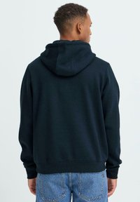 Felpa con cappuccio blu navy con coulisse e polsini a coste, realizzata in tessuto morbido. Indossata con jeans di denim azzurro chiaro.