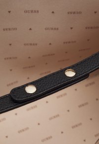 Sort tekstureret kunstlæderrem med guldhardware på et blødt beige interiør med et gentaget "GUESS" logo mønster.