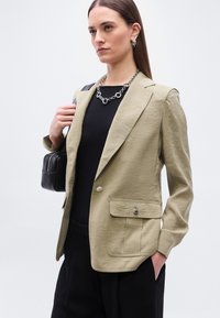 Γυναίκα με μπεζ σακάκι, μαύρο τοπ και ασημένιο κολιέ με αλυσίδα, κρατώντας μαύρη τσάντα στον ώμο, σε απλό φόντο.