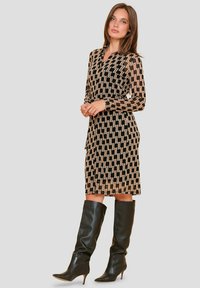 Zwart en beige jurk met zigzagpatroon, lange mouwen en V-hals. Boven de knie lengte, gecombineerd met kniehoge zwarte leren laarzen.