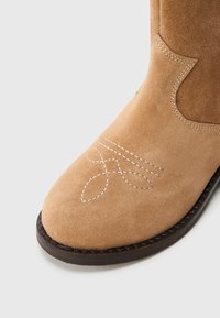 Stivaletto in suede color nocciola con punta rotonda e un motivo decorativo di cuciture bianche. La suola è scura e testurizzata, aumentando la durabilità.