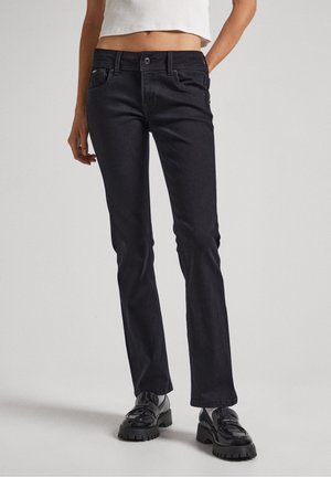 Kobieta ubrana w czarne obcisłe dżinsy, biały krótki top i czarne masywne mokasyny, stojąca na tle jednolitego jasnoszarego tła.