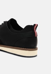 Baskets noires avec talon perforé, col de cheville tricoté, languette de talon en tissu rayé rouge et blanc, semelle superposée crème et marron.