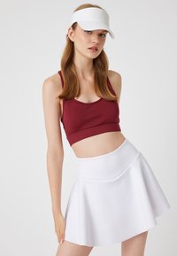 Maroon urheiluliivit yhdistetty valkoiseen skeittihameeseen. Asusteessa on sileä kangas, ja se on istuva ja kellotettu muotoilultaan. Mukana myös valkoinen lippalakki.