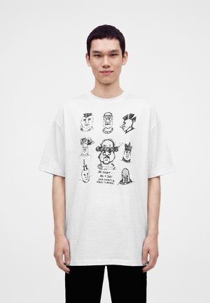 Νέος άνδρας που φοράει λευκό φαρδύ T-shirt με αφηρημένα μαύρα σκίτσα προσώπου και χειρόγραφο κείμενο μπροστά, στέκεται μπροστά σε απλό φόντο.