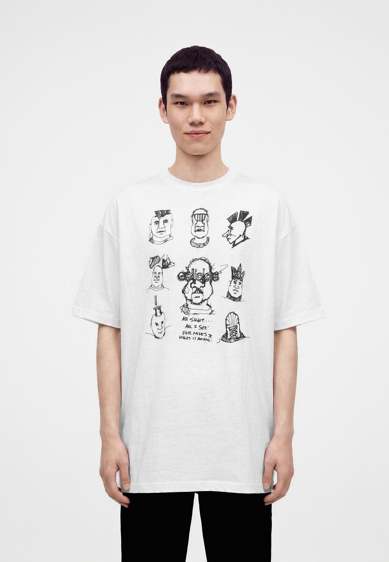 Νέος άνδρας που φοράει λευκό φαρδύ T-shirt με αφηρημένα μαύρα σκίτσα προσώπου και χειρόγραφο κείμενο μπροστά, στέκεται μπροστά σε απλό φόντο.