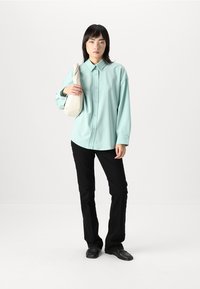 Camicia button-up azzurro chiaro in tessuto morbido, abbinata a pantaloni neri slim-fit e scarpe nere. Tieni in mano una borsa color crema.