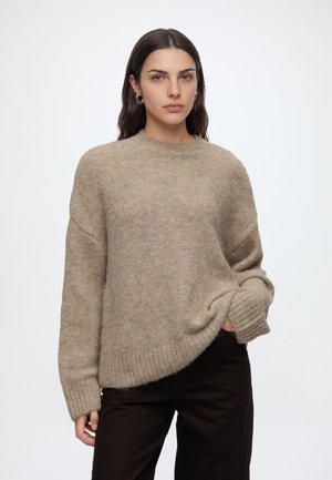 Vrouw met lang donker haar met gouden creolen, een losse beige gebreide trui en een zwarte broek tegen een effen lichte achtergrond.