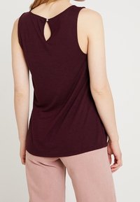 Bordeauxfärgad ärmlös topp med en nyckelhålsöppning bak. Mjuk, lätt väv med en avslappnad passform, som visas ihop med ljusrosa byxor.