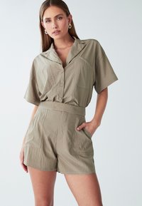 Moteris su smėlio spalvos marškiniais su mygtukais ir atvartais, dėvinti derančias šortus iki juosmens, viena ranka kišenėje, prieš paprastą šviesų foną.