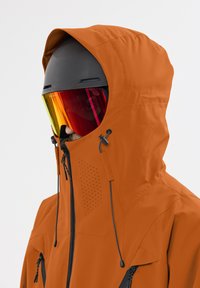 Orangen wasserdichte Jacke mit Kapuze, ausgestattet mit Reißverschluss-Taschen, Belüftungsöffnungen und einem hochkragens Design. Tragen mit einem schwarzen Helm und reflektierendem Visier.