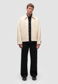 Casualna kurtka polarowa w kolorze ecru; prosty krój z kołnierzykiem, dwoma przednimi kieszeniami i czarnym zapięciem na guziki; noszona na czarną koszulę.