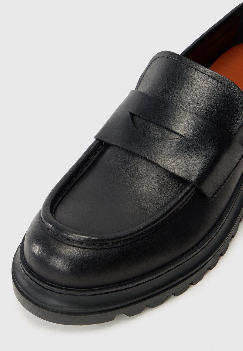 Sorte læder loafers med en glat tekstur, en syet kant og et polstret bånd over toppen. Tykk, ribbet gummisål.