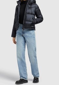 Personne portant une doudoune noire, un col roulé noir, un jean droit bleu clair et des bottines noires sur un fond uni.