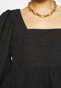 Svart texturerad klänning med fyrkantig halsringning, puffärmar och utsvängd kjol. Bärs med ett kraftigt halsband i guld.