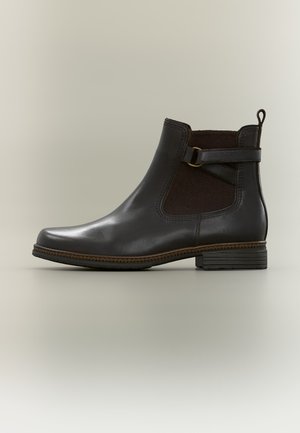 Bottine en cuir noir avec panneau élastique latéral, sangle décorative et petit talon empilé sur fond gris clair.