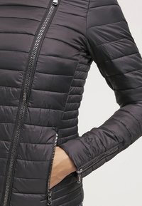 Chaqueta negra acolchada con textura acolchada horizontal, dos bolsillos con cremallera y puños con cremallera. Presenta un acabado liso y brillante.