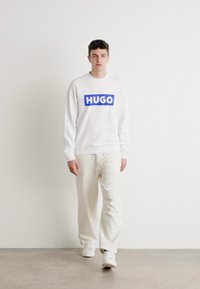 Baltas džemperis su mėlynu "HUGO" logotipu, suderintas su šviesiai smėlinėmis kelnėmis ir baltais sportbačiais, pateiktas prieš neutralų foną.