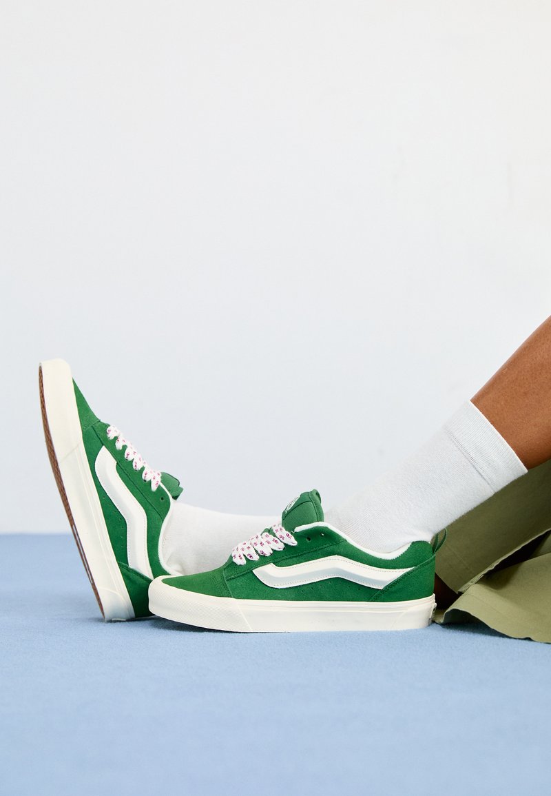 Baskets en daim vert avec des accents blancs, lacets texturés avec des détails roses et une semelle en caoutchouc beige, portées avec des chaussettes blanches et un short vert.