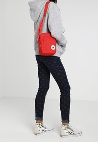 Osoba v šedé mikině s kapucí, tmavých vzorovaných legínách, kostkovaných vysokých teniskách a s malou červenou kabelkou přes rameno s kulatou nášivkou.