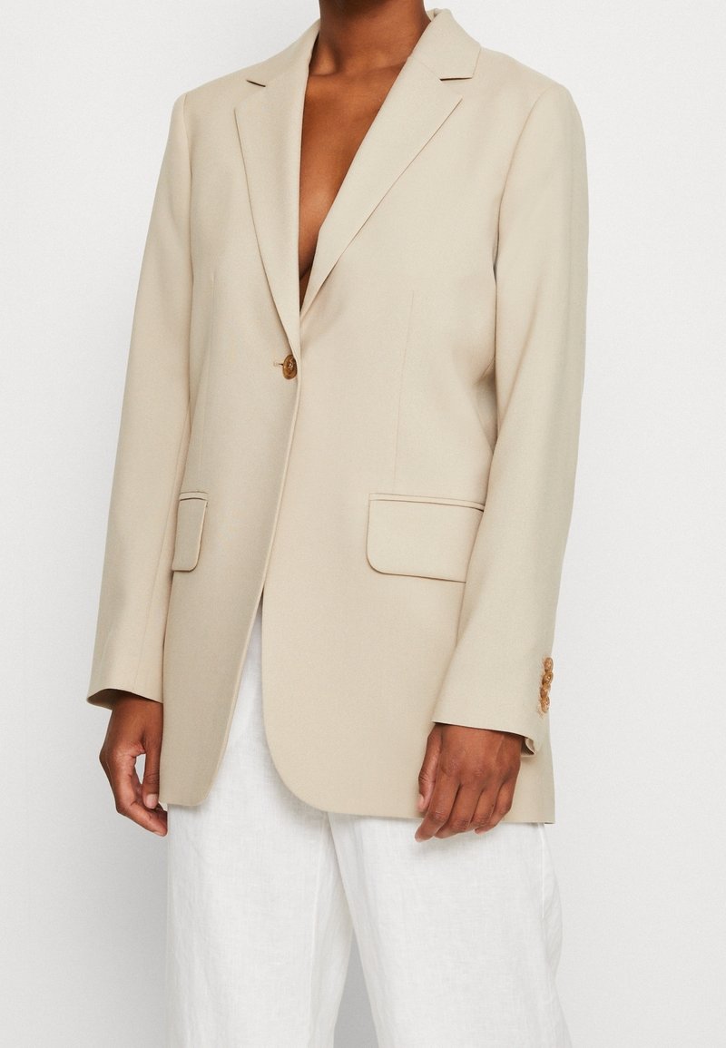 Blazer beige à un seul bouton avec revers crantés et poches à rabat, porté sur un pantalon blanc par une personne à la peau foncée.
