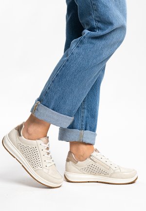 Light beige sneakers with perforated sides, smooth matte texture, and metallic accents, paired with cuffed blue denim jeans.