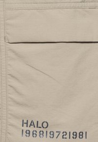 Beige stoffen zak met klep en gestikte randen, met donkere sjabloonstijl tekst "HALO 1968 1972 1981" nabij linksonder.
