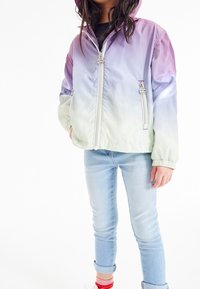 Letvægts bomberjakke i gradient fra lilla til grøn, med lynlås, hætte, elastiske manchetter og to sidelommer. Båret med blå jeans og røde sokker.