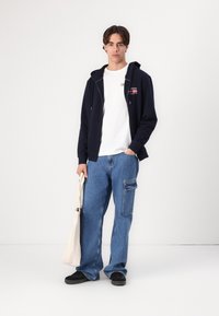 Hoodie marine avec fermeture éclair, T-shirt blanc et pantalon cargo en denim baggy bleu. Le mannequin tient un sac fourre-tout beige et porte des baskets noires.