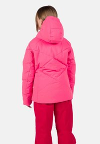 Giacca imbottita rosa con cappuccio, caratterizzata da un design trapuntato. Indossata con pantaloni rosa coordinati, che mettono in risalto una finitura liscia e brillante.