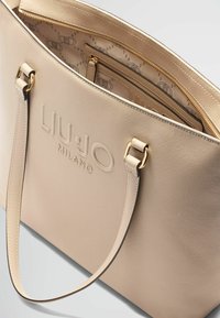 Sac fourre-tout beige en matériau synthétique texturé, logo en relief "LIU.JO MILANO", quincaillerie dorée et compartiments zippés.