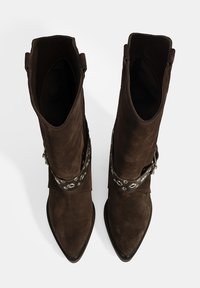 Braune Wildleder-Stiefeletten mit spitzer Zehenform, die mit einem dekorativen Riemen mit silbernen Ösen und einer strukturierten Oberfläche versehen sind.