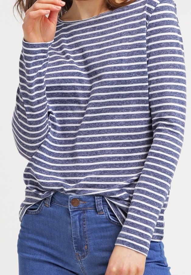 Femme portant un t-shirt à manches longues rayé horizontalement bleu marine et blanc, légèrement rentré dans un jean bleu, main près du visage.