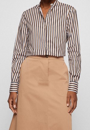 Osoba nosząca koszulę z długim rękawem w pionowe paski w kolorze białym, brązowym i czarnym, połączoną z beżową spódnicą z wysokim stanem zapinaną na guziki.