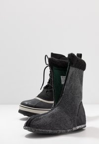Schwarze Lederwinterstiefel mit weißer Sohle und Schnürsenkeln neben grauen Filzstiefeln mit schwarzen und grünen Akzenten sowie sichtbaren Nähten.