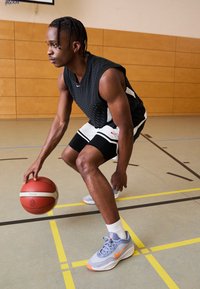 Basketballspiller i sort nettrøje og sorte shorts, der dribler med en bold. Bærer lysegrå Nike-sko med orange detaljer og hvide strømper.