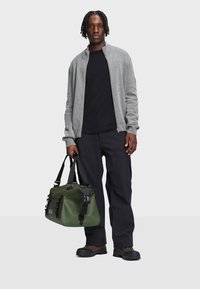 Jonge man met korte dreadlocks, gekleed in een grijze ritscardigan, zwart shirt, zwarte broek, bruine schoenen, en houdt een groene sporttas vast.