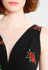 Kobieta w czarnej bluzce bez rękawów z czerwoną haftowaną różą, eksponująca szyję, ramiona i linię żuchwy na jednolitym jasnym tle.