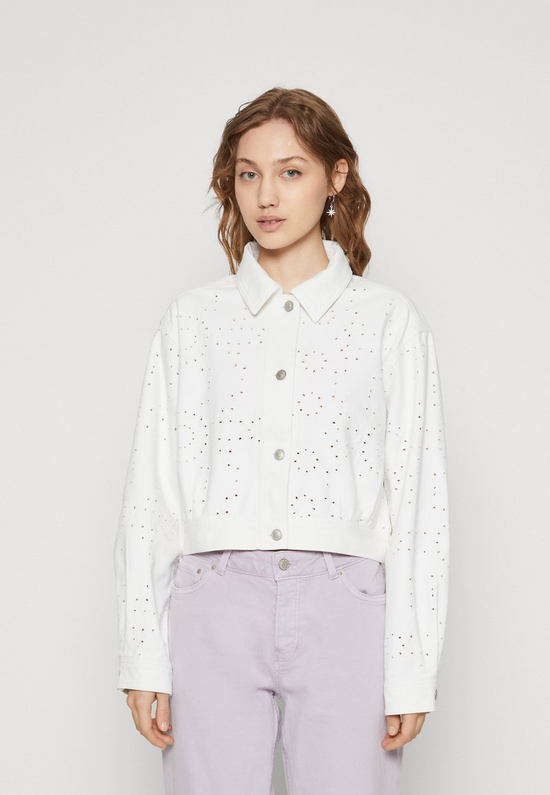 Chaqueta blanca cortada con un cierre de botones en la parte delantera, que presenta agujeros cortados con láser y un cuello estructurado. Combinada con pantalones de color púrpura claro.